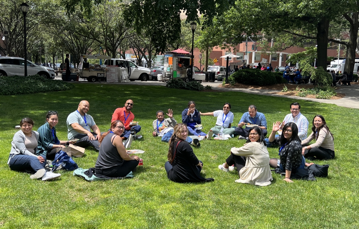 A group of 14 fellows sit for a picnic lunch on s sunny day in the Harkness yard and Yale School of Medicine